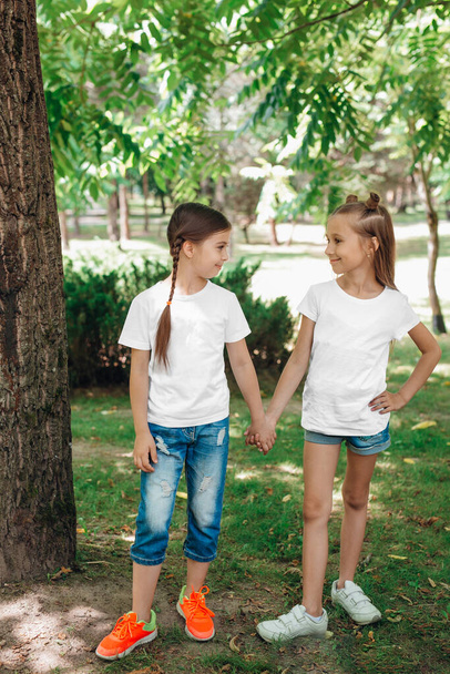 Two little girls in white t-shirts stand holding hands in park outdoor. Mock up. - Φωτογραφία, εικόνα