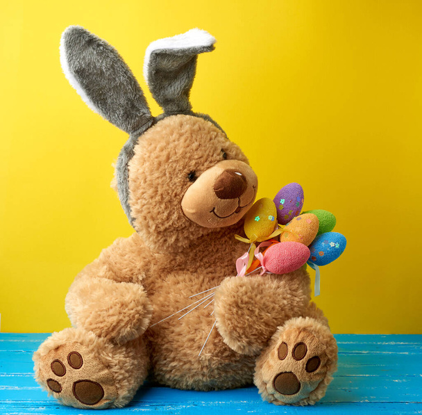 grote schattige bruine teddybeer met kleurrijke paaseieren, het dragen van een konijn masker met lange oren op zijn hoofd, grappige vakantie kaart - Foto, afbeelding