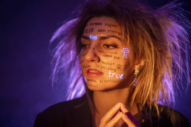 Girl with Disheveled Hair in Dramatic Purple light with written different language words on her face. Beautiful model with UV Fluorescent neon creative make-up. face close up - Photo, Image