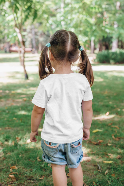 back views of little girl in blank t-shirt in park. Mockup for design - Фото, изображение