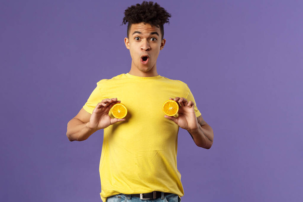 Holidays, vitamins and vacation concept. Portrait of funny and cute young 25s man fool around, showing breast with pieces of oranges over chest, look ashamed or shocked, purple background - Photo, Image
