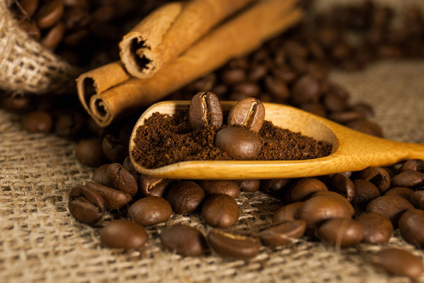 Primer plano, cucharada de madera con café molido y granos de café, palitos de canela sobre un fondo de textura de arpillera
 - Foto, imagen