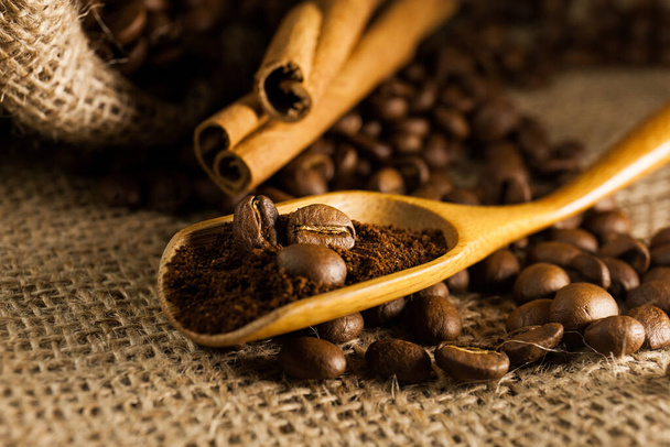 Close-up, houten schep met gemalen koffie en koffiebonen, kaneelstokjes op een jute getextureerde achtergrond - Foto, afbeelding