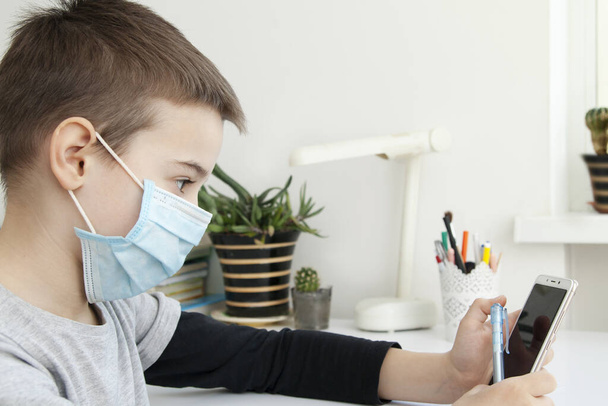 Schoolboy with a smartphone at the desktop with lessons. Coronavirus Quarantine Distance Learning Concept - Photo, Image