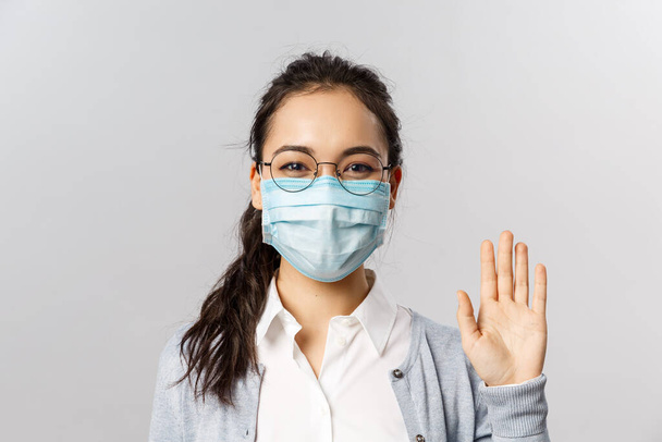 Covid19, vírus, egészség és orvostudomány. Portré barátságos fiatal lány orvosi maszk mond hello, hello integető kéz és vigyorgó szem, vigyázz pandémia alatt, karantén - Fotó, kép