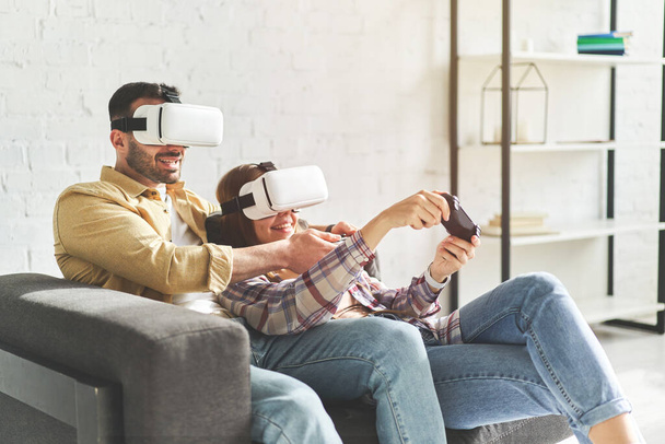 Νεαρό χαμογελαστό ζευγάρι με γυαλιά εικονικής πραγματικότητας που παίζει βιντεοπαιχνίδι στον καναπέ - Φωτογραφία, εικόνα