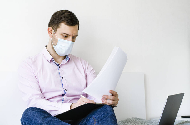 Coronavirus. Quarantine. A man in a shirt and jeans solves work issues remotely. In the mask. Documentation. Laptop and phone. Coronavirus pandemic in the world. - Фото, изображение