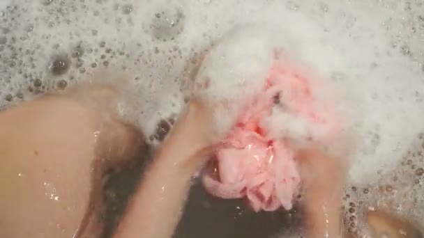 Female hand holds pink washcloth with soap foam at bath - Imágenes, Vídeo