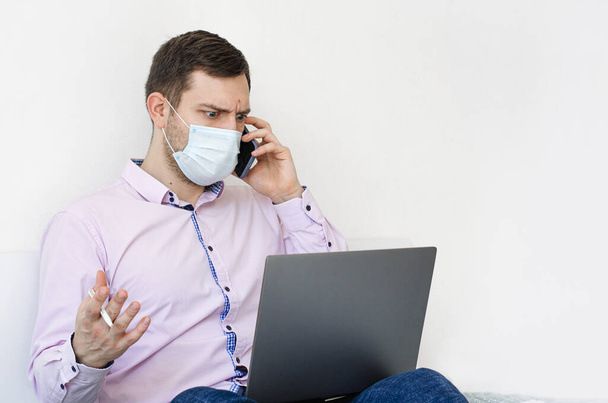 Coronavirus. Quarantine. A man in a shirt and jeans solves work issues remotely. In the mask. Documentation. Laptop and phone. Coronavirus pandemic in the world. - Foto, afbeelding