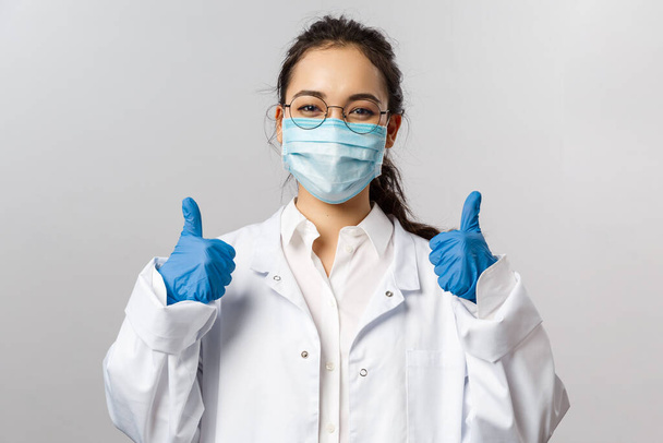 医師、感染症、研究とcovid19の概念。満足のいく若いアジアの女性医師は、ウイルスの研究、ワクチンの発見、親指アップを示す、顔のマスクと手袋に良い結果を受け取ります - 写真・画像