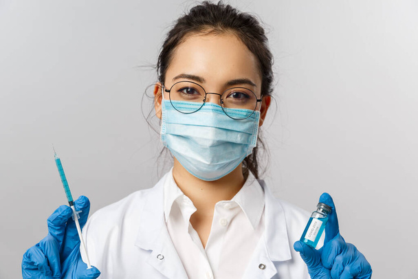 Ärzte, Infektiologen, Forschung und Covid19 Konzept. Nahaufnahme einer asiatischen Krankenschwester in medizinischer Maske, Handschuhen und weißem Mantel, die Spritze und Ampulle mit Coronavirus-Impfstoff in der Hand hält, heilt Menschen - Foto, Bild