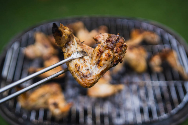 夏の週末に焼く。焼きたての肉。鶏の羽をトングのペアで持ち上げる。うまい焼き肉の盛り合わせ. - 写真・画像