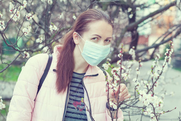 Jonge vrouw met een masker staat in de buurt van een bloeiende boom op straat. - Foto, afbeelding