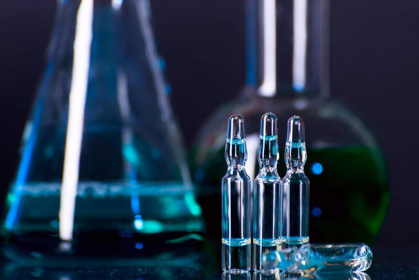 Glass ampoules with liquid for injection against lab flasks in dark laboratory. COVID-19 background. - Photo, Image