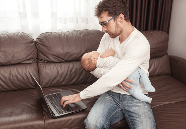Young father working on the laptop at home with a little baby on his hands. Self isolation. Freelancer.  - Фото, изображение