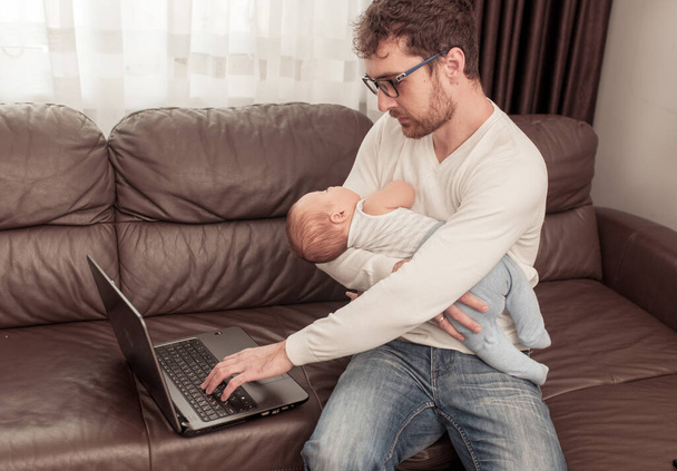 Father working at home using a laptop with a little baby on his hands. Freelancer. Stay at home concept. - 写真・画像