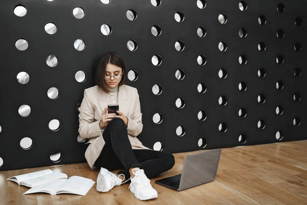 Education, people and university concept. Portrait of young smart girl studying in business school, learning in coworking space, sitting on floor with laptop and mobile phone, with homework - Foto, immagini
