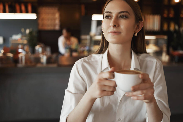 Satisfied relaxed young lady, businesswoman drinking coffee at cafe, looking out window enjoying nice cozy atmosphere, having office break, smiling thoughtfully, leisure and business concept - Photo, image