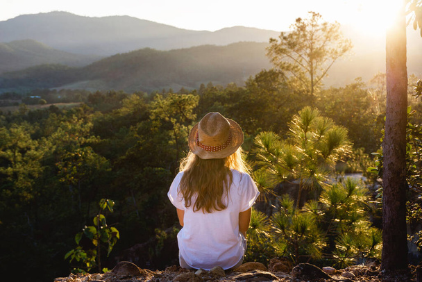 Young girl sitting on the edge of the cliff watching sunset. Beautiful forest and hills landscape. Travel lifestyle background. - Foto, afbeelding