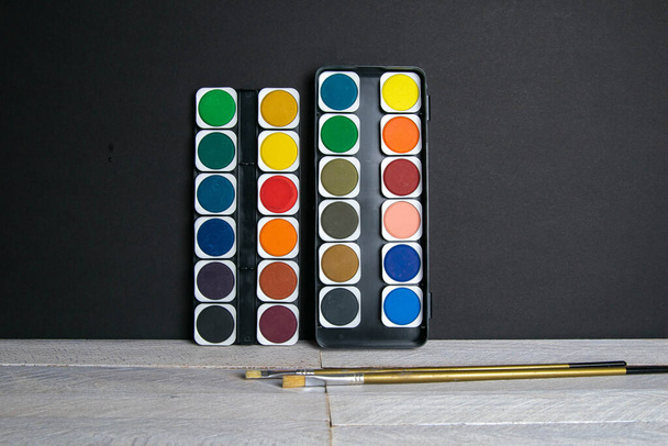 Bunte Farben stehen auf einem Holztisch vor einer schwarzen Wand. Frontalansicht. - Foto, Bild