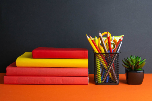 A stack of books, a green flower, and a glass of pencils stand on an orange background. - Фото, изображение