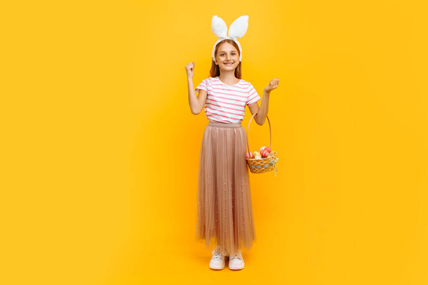 Pełna długość, mała atrakcyjna dziewczynka na głowie z króliczymi uszami, trzymająca koszyk pisanek i pokazująca gest zwycięstwa na żółtym tle. Symbol Wielkanocy i wiosny - Zdjęcie, obraz