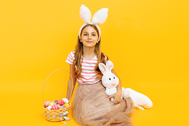 A happy beautiful girl on a head with rabbit ears holds a wicker basket with colorful eggs and an Easter Bunny on a yellow background. Symbol of Easter and spring - Photo, image