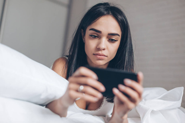Serious Beautiful woman using smartphone while lying on her bed. Smiling Brunette Girl holding mobile phone - Photo, image