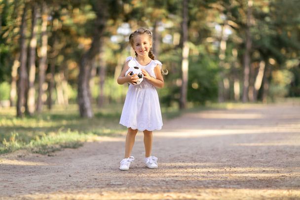 Πορτρέτο ενός χαμογελαστού κοριτσιού με μια μπάλα ποδοσφαίρου στα χέρια της. Στο πάρκο στο δρόμο - Φωτογραφία, εικόνα