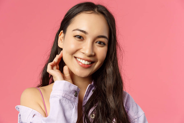 Close-up portrait of tender, beautiful young asian woman with clean skin, no pimples or blemishes, smiling lovely camera, touching neck and look camera romantic, stand pink background - Photo, Image