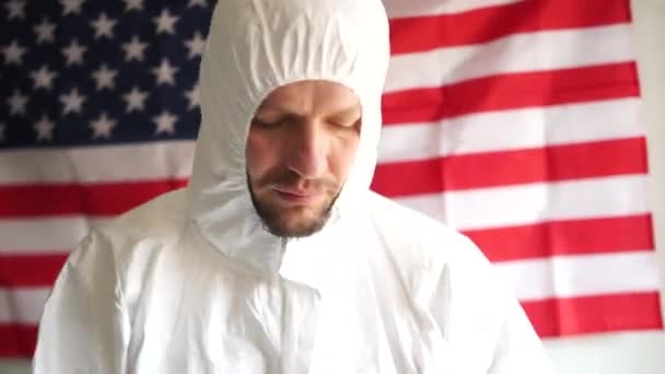 Male doctor in a protective suit puts on a mask and glasses on the background of the usa flag. Coronovirus epidemic in the United States of America - Footage, Video