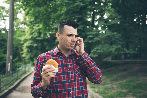 Handsome young man eating sandwich autdoor. He is holding a phone - Photo, image