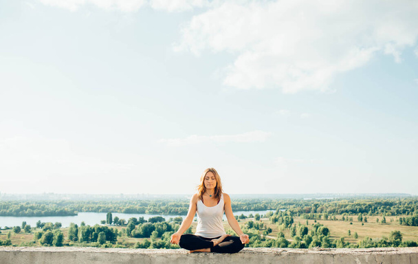 Young woman practices yoga outside. Calm peaceful girl sitting on parapet in lotus position. Her hands lowered to knees. Trees river and sky on background. - Fotografie, Obrázek