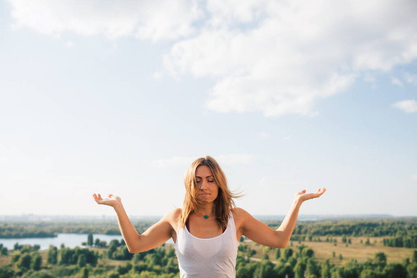 Young woman practices yoga outside. Calm blonde girl with closed eyes. She dressed in white T-shirt. Her palms turned to sky. Portrait. Trees river and sky on background. - Photo, Image