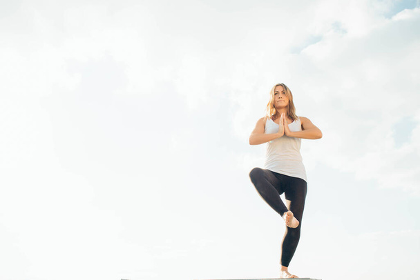 Young woman practices yoga outside. Blonde girl standing on one leg, another raised bent at knee. Hands at chest level, palms touching. Sky and clouds on background. - Foto, Imagem