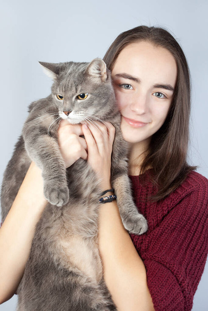 Güzel gülümseyen esmer kollarında büyük çizgili safkan gri bir kedi tutuyor. - Fotoğraf, Görsel