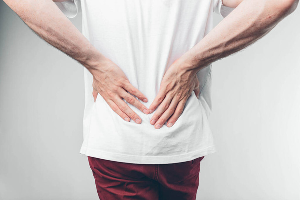 Caucasian man in white T-shirt and burgundy pants. His hands holding on lower back. Cut view. Concept. Loins hurts. Isolated over light background. - Photo, Image