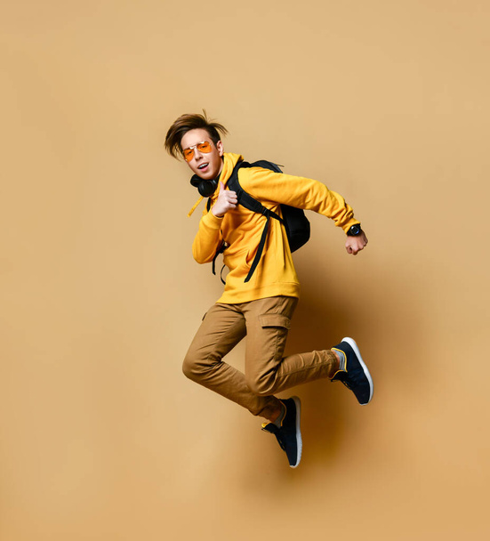 Young teen boy in comfortable clothing, sneakers and backpack jumping over yellow wall background - Fotó, kép