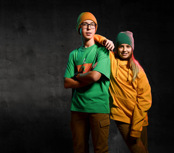 Young happy teens boy and girl in green and yellow stylish casual clothing and hats standing and smiling over dark background - Φωτογραφία, εικόνα