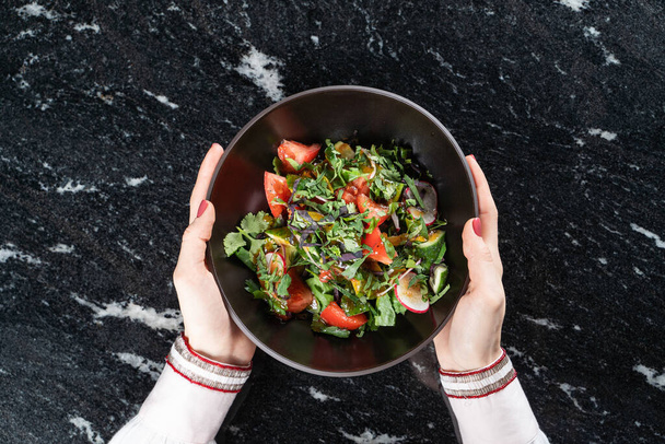 Vrouw houdt een kom met groentesalade met handen, boven zwart marmeren oppervlak, direct boven zicht - Foto, afbeelding