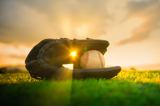 Honkbal in de handschoen in het gazon bij zonsondergang in de avond dag met zonnestralen en lens flare licht - Foto, afbeelding