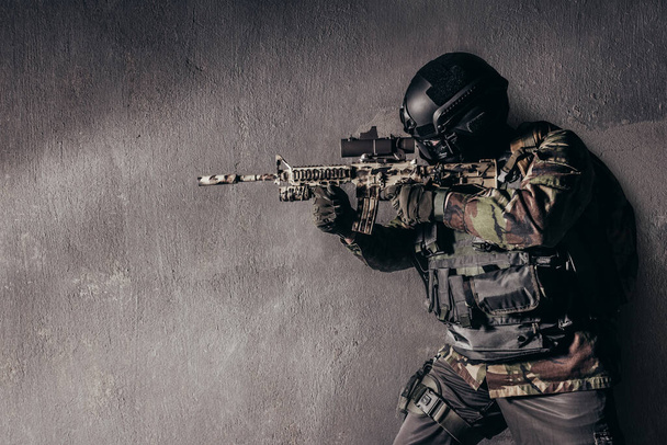 Kamuflaj üniformalı tam teçhizatlı bir askerin kirli beton arka planda duran ve nişan alan bir tüfeğin fotoğrafı.. - Fotoğraf, Görsel