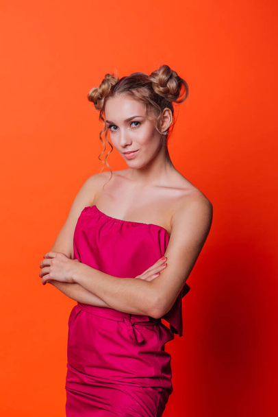 The girl in a red dress on a orange background in the studio. Blonde girl with two hair knots holding hands on chest, looking to the camera and smiling. - Foto, Bild