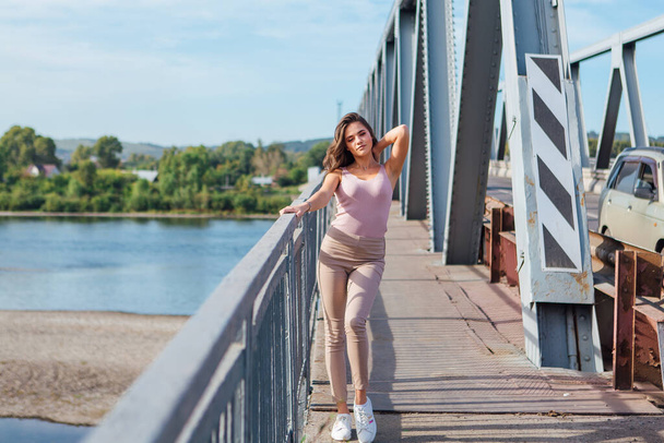 Warm summer portrait of a young beautiful woman posing on the old rusty transport bridge over the river during sunset. - 写真・画像