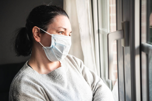 Beteg nő maszkkal és orvosi kesztyűvel, aki az ablakon néz be, valószínűleg megfertőződött a koronavírussal és izolálta magát. - Fotó, kép