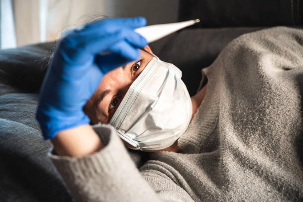 Chora kobieta w masce i rękawiczkach medycznych patrząca na termometr leżący na sofie, prawdopodobnie zarażona koronawirusem. - Zdjęcie, obraz