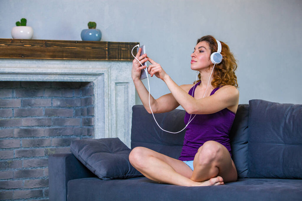 Nice woman in a purple top is sitting on the sofa and choosing music to listen with headphones - Foto, afbeelding
