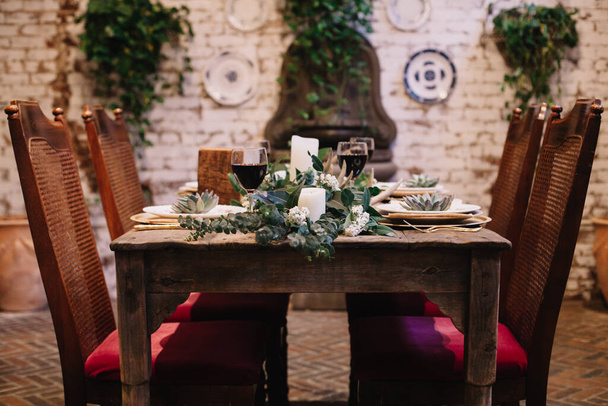 Rusztikus esküvői asztaldíszítés vintage tányérokkal, zöld-fehér eukaliptusz koszorúval, szarvasagancsokkal, arany edényekkel. Boho stílusban. Asztalkészlet egy privát vacsorára, eseményre, partira, dátumra vagy esküvőre. Felszolgált vörösbor - Fotó, kép