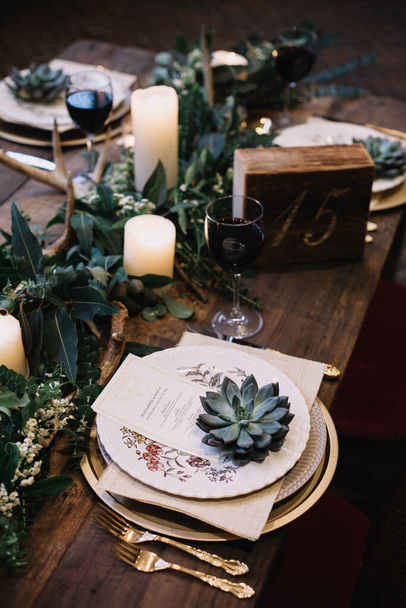 Сельский свадебный стол со старинными тарелками, зелеными и белыми эвкалиптовыми гирляндами, оленьими рогами, золотой посудой. В стиле Бохо. Стол накрыт на событие, вечеринку, дату или свадьбу. Красное вино подается
 - Фото, изображение