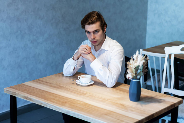 Hombre guapo pensativo en camisa blanca mirando hacia otro lado sentado en la mesa en la cafetería pensando en los planes, chico caucásico esperando para reunirse en la cafetería soñando con la idea de ocio
 - Foto, imagen